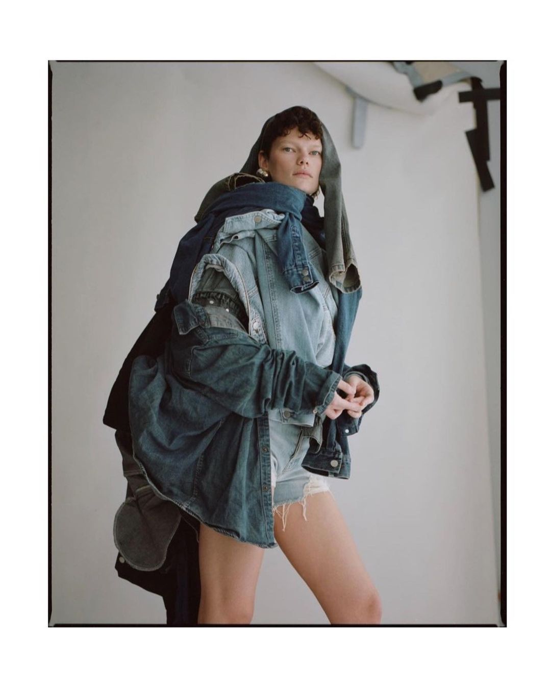 Woman posing in a photo studio wearing a denim outfit made of different denim jackets and shorts, against a white backdrop.
