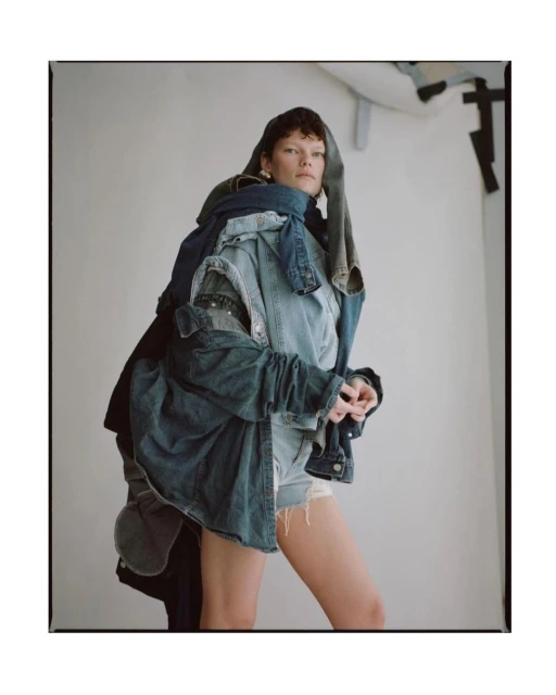 Woman posing in a photo studio wearing a denim outfit made of different denim jackets and shorts, against a white backdrop.