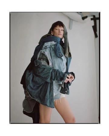 Woman posing in a photo studio wearing a denim outfit made of different denim jackets and shorts, against a white backdrop.
