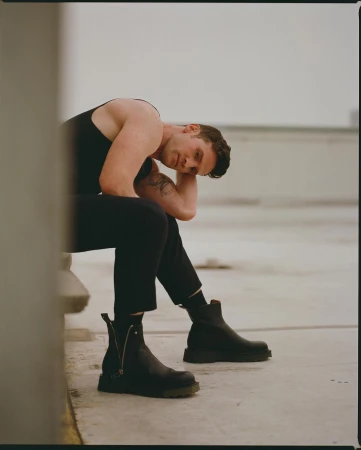 Adult male in tank top and pants sits on a concrete structure in black boots, with a building and grey sky in the background.