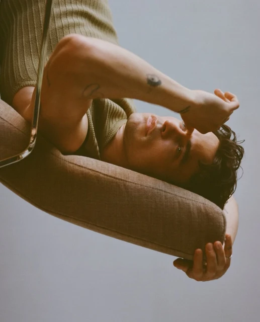 An adult male reclines in a tan chair against a soft grey background, with his arm raised to his face in a casual, relaxed pose.