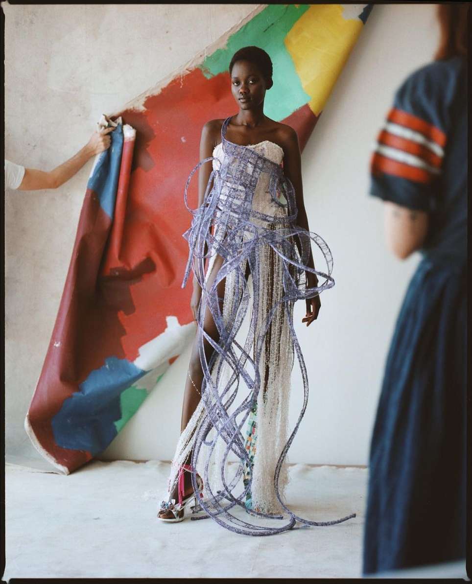 A slender Black woman in a white and blue dress poses indoors, with two people partially visible holding a colorful canvas.