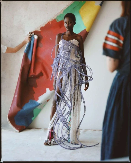 A slender Black woman in a white and blue dress poses indoors, with two people partially visible holding a colorful canvas.