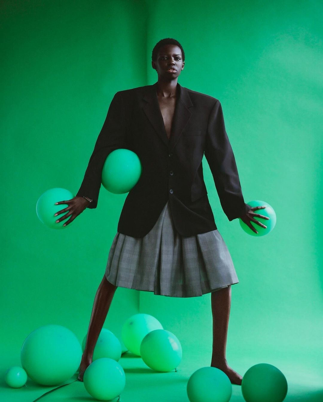 Full-length shot of a Black female in a black blazer, gray skirt, surrounded by green balloons against a vibrant green backdrop.