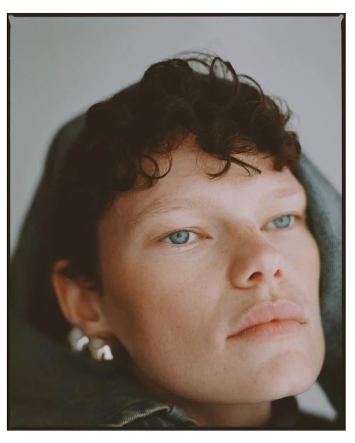 Close-up portrait of a young adult woman with short curly brown hair, wearing silver earrings and a green jacket against a grey background.