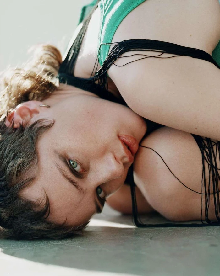 Close-up portrait of a woman with short brown hair and green eyes lying on a light grey surface, wearing a green top and black straps.