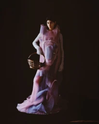 Model in voluminous pink and lilac textured dress holding a basket against a dark background