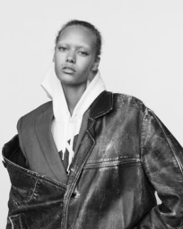 Monochrome portrait of a model wearing a heavy leather jacket over a layered hoodie and blazer against a plain white background.
