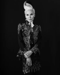 Monochrome portrait of a woman with a high sculpted hairstyle wearing an embroidered jacket and a metallic neckpiece.