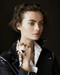 Portrait of a woman with dark short hair wearing a dark jacket and white shirt, featuring a playful character charm bracelet.