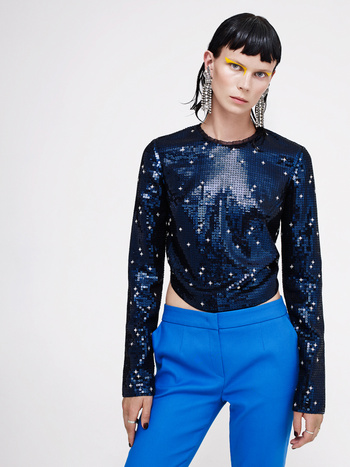 Fashionable adult woman in a sequined top and bright blue pants against a white backdrop, showcasing a modern and stylish clothing ensemble.