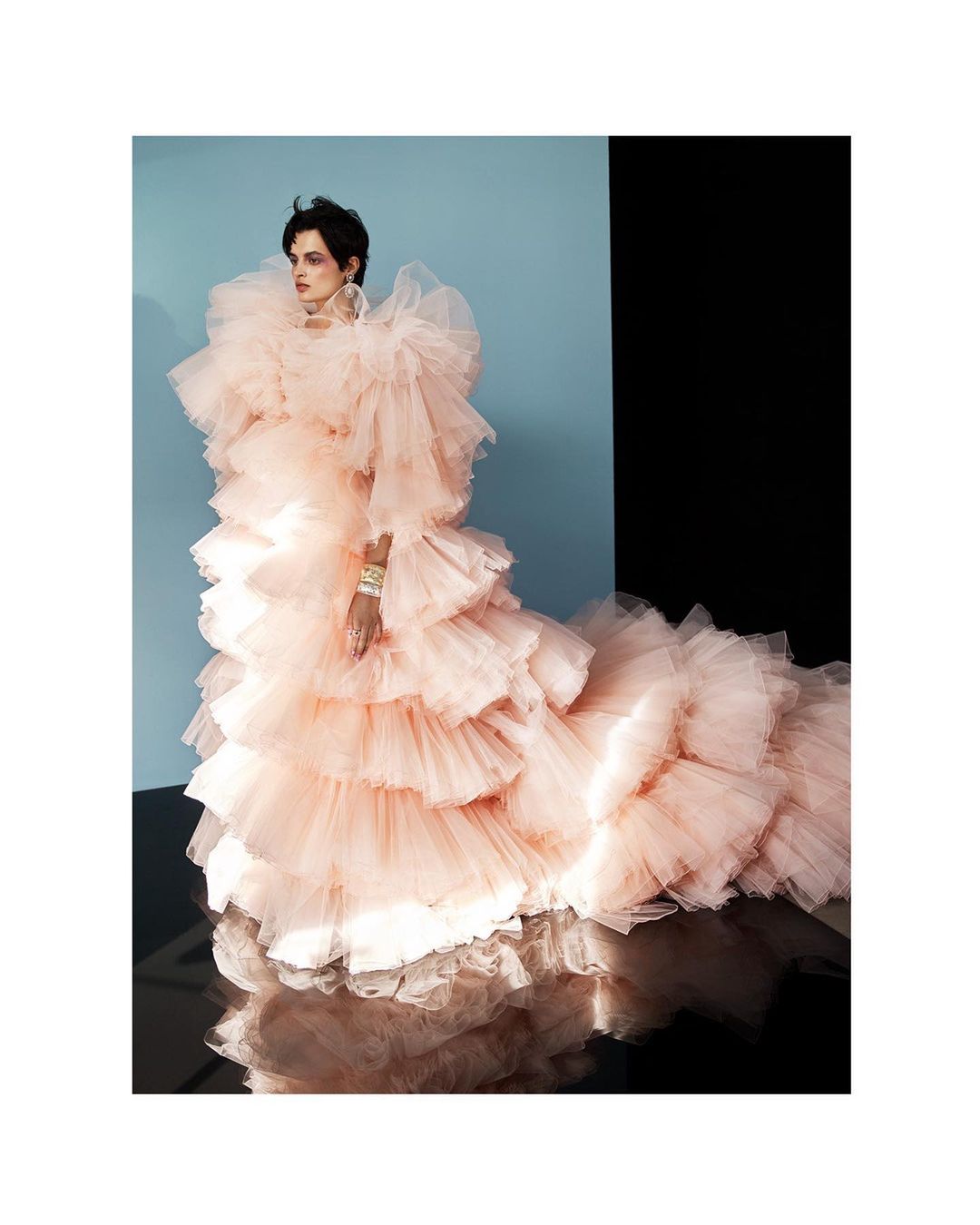 Woman in a voluminous ruffled pale pink gown with an elaborate train poses in a photo studio against blue and black backgrounds.