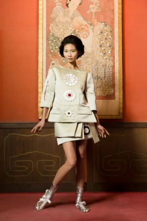 Model in off-white structured dress and silver boots standing against a red wall with traditional artwork