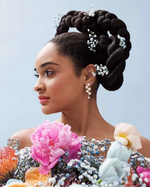 Portrait of a woman in profile with an elaborate braided hairstyle adorned with baby's breath, wearing pearl earrings and floral top.