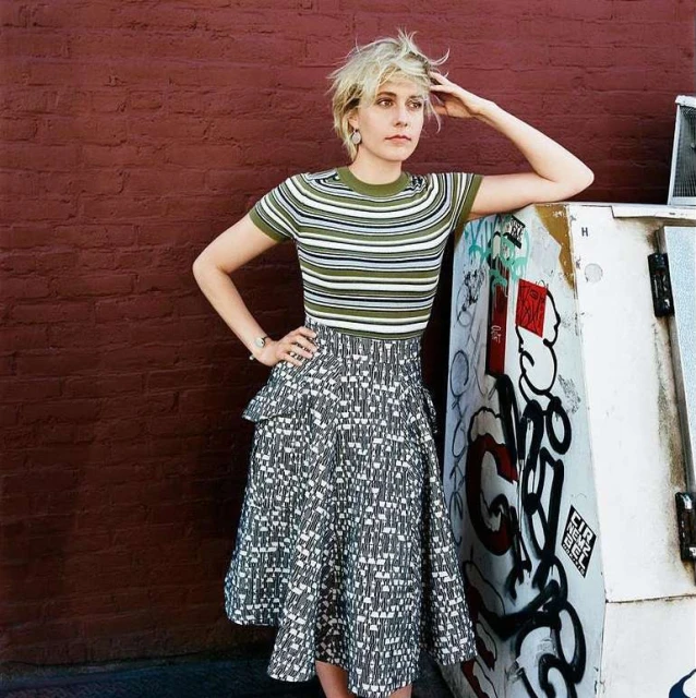 Blonde woman in striped top and patterned skirt poses outdoors against a red brick wall and graffiti-covered utility box.