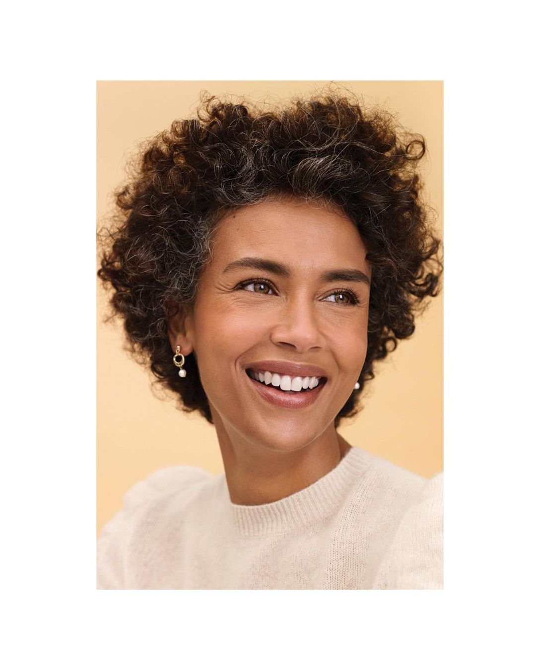 Close-up of a woman with curly hair smiling and wearing pearl earrings and a cream-colored sweater against a peach background.
