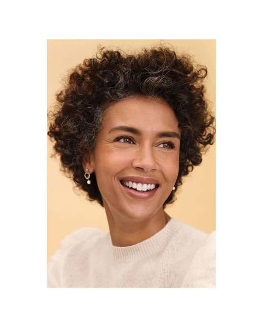 Close-up of a woman with curly hair smiling and wearing pearl earrings and a cream-colored sweater against a peach background.