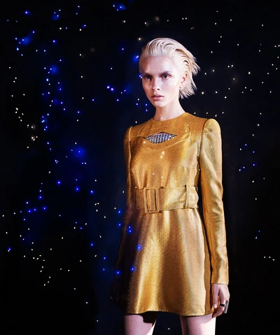 Young woman with bleached hair poses in a gold mini dress with a belt against a starfield background for a fashion or beauty editorial.