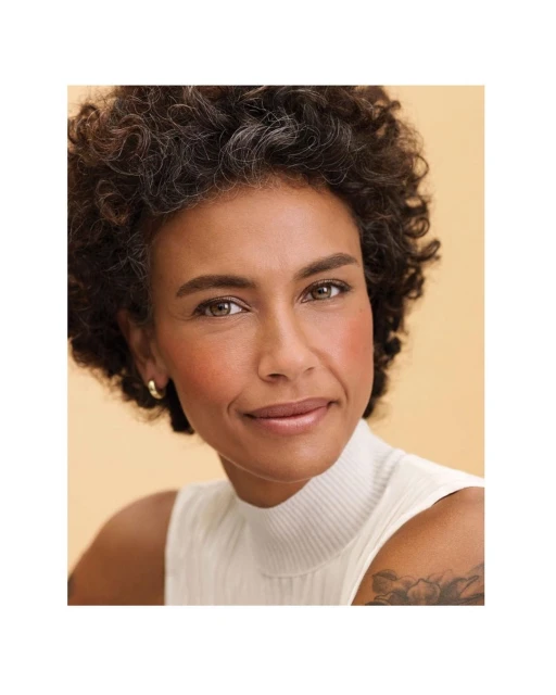 Close-up portrait of adult woman with curly hair and fair skin wearing a white top against a light beige background in studio lighting.