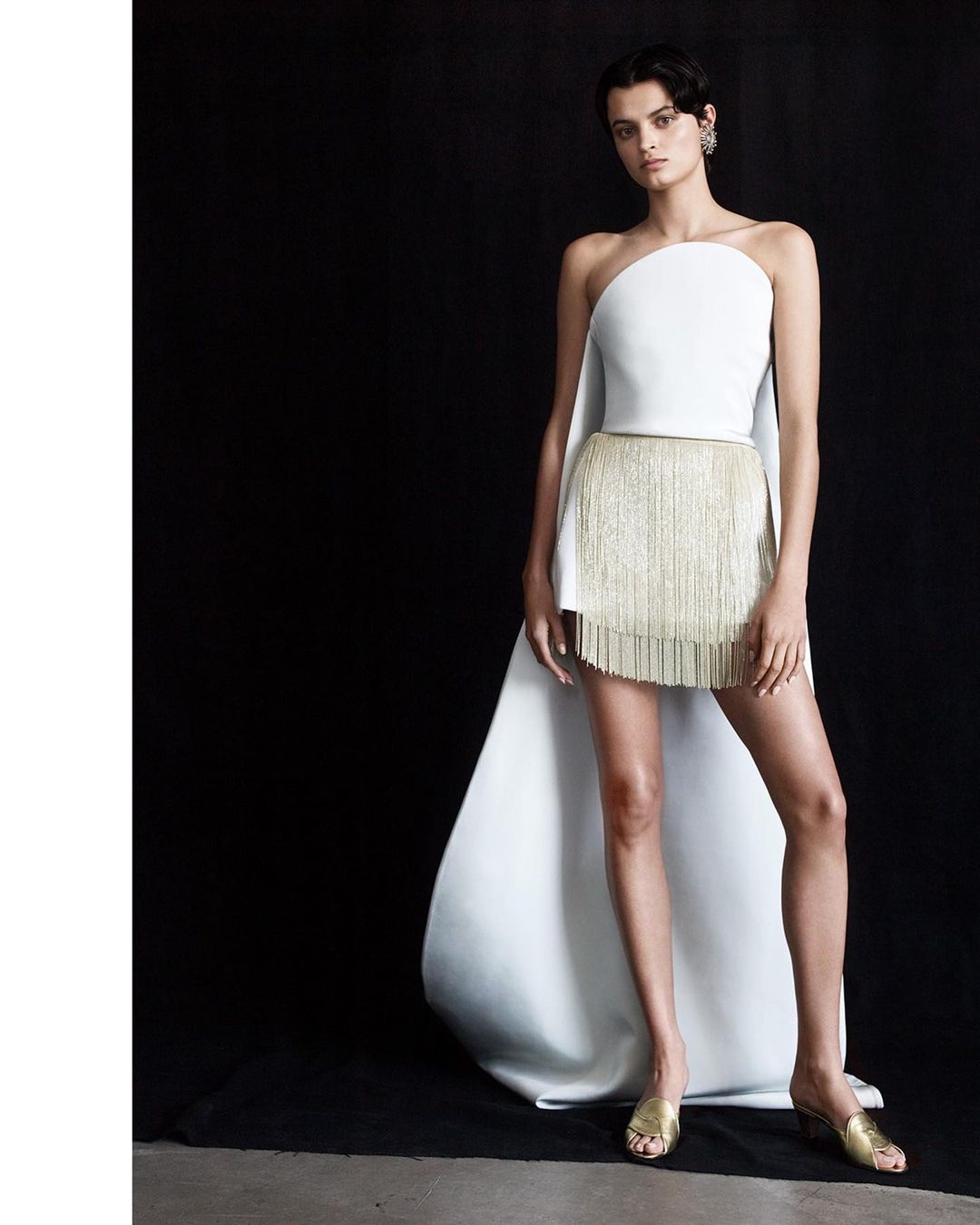 Fashion model poses in a white strapless top with a gold fringe skirt and white flowing cape against a dark background in a studio shot.