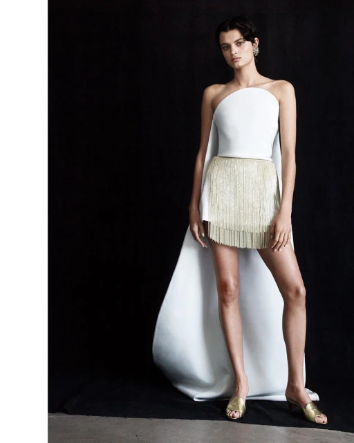 Fashion model poses in a white strapless top with a gold fringe skirt and white flowing cape against a dark background in a studio shot.