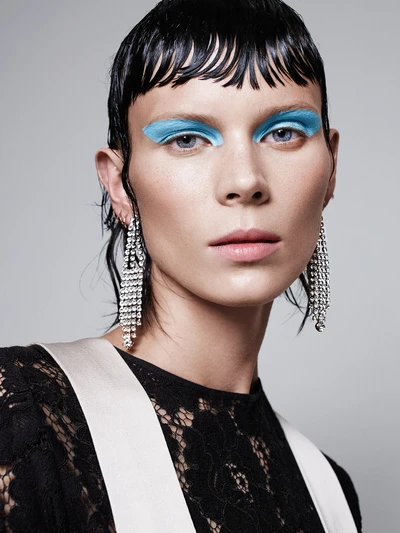 Close-up portrait of a woman with blue eyeshadow, cascading rhinestone earrings, and black lace top against a grey background.