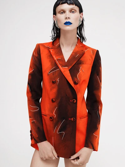 Adult woman with blue lipstick wearing an orange printed blazer in a studio setting, jewelry earrings, and rings on her fingers.