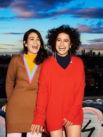 Two smiling women stand outdoors on a rooftop at dusk with city skyline. One woman wears a brown sweater dress, the other wears a red sweater.