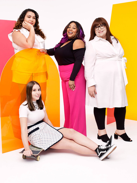 Four diverse women in a studio setting. One woman seated on a skateboard, dressed in white and black, with colorful backdrop panels.