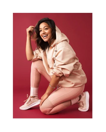 Smiling woman in pink hoodie and matching leggings crouching against a deep red studio backdrop