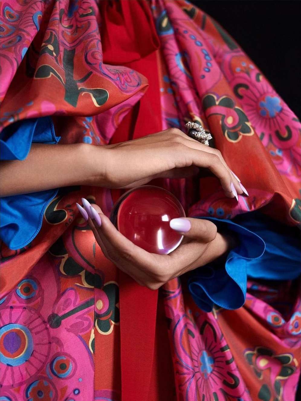 Close-up shot of hands holding a crystal ball, vibrant patterned fabric background with shades of pink, red, and blue.