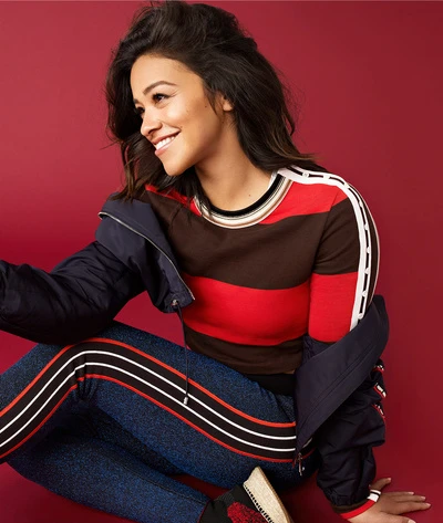Smiling adult woman with long brown hair poses in an indoor studio, wearing a striped shirt, jeans, and a jacket. Portrait. 