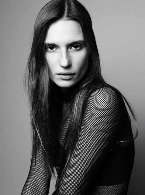 Monochrome portrait of a young adult female with long brown hair, wearing a black mesh top, posing in a photo studio setting.