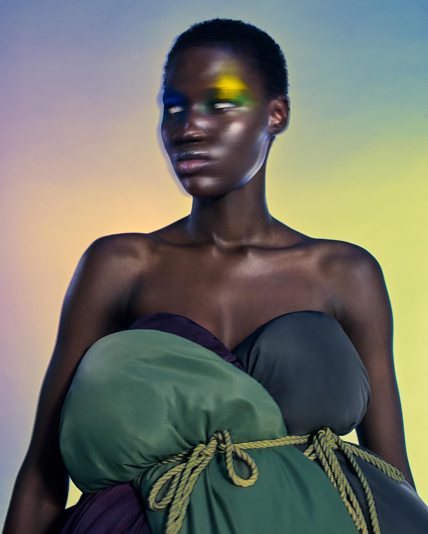 Striking studio shot of a dark-skinned woman with colorful eyeshadow and a unique garment made of green, black, and purple fabric bundles.