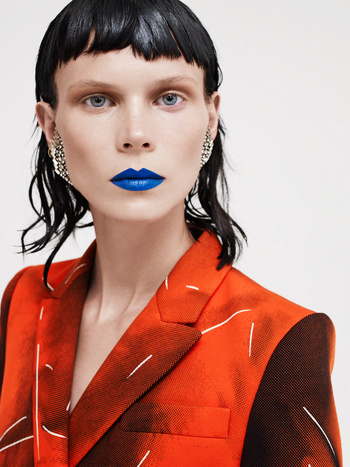 Fashion portrait of a woman with blue lipstick wearing an orange patterned blazer against a white background in a studio setting.