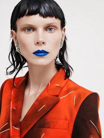 Fashion portrait of a woman with blue lipstick wearing an orange patterned blazer against a white background in a studio setting.