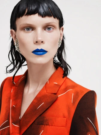 Fashion portrait of a woman with blue lipstick wearing an orange patterned blazer against a white background in a studio setting.