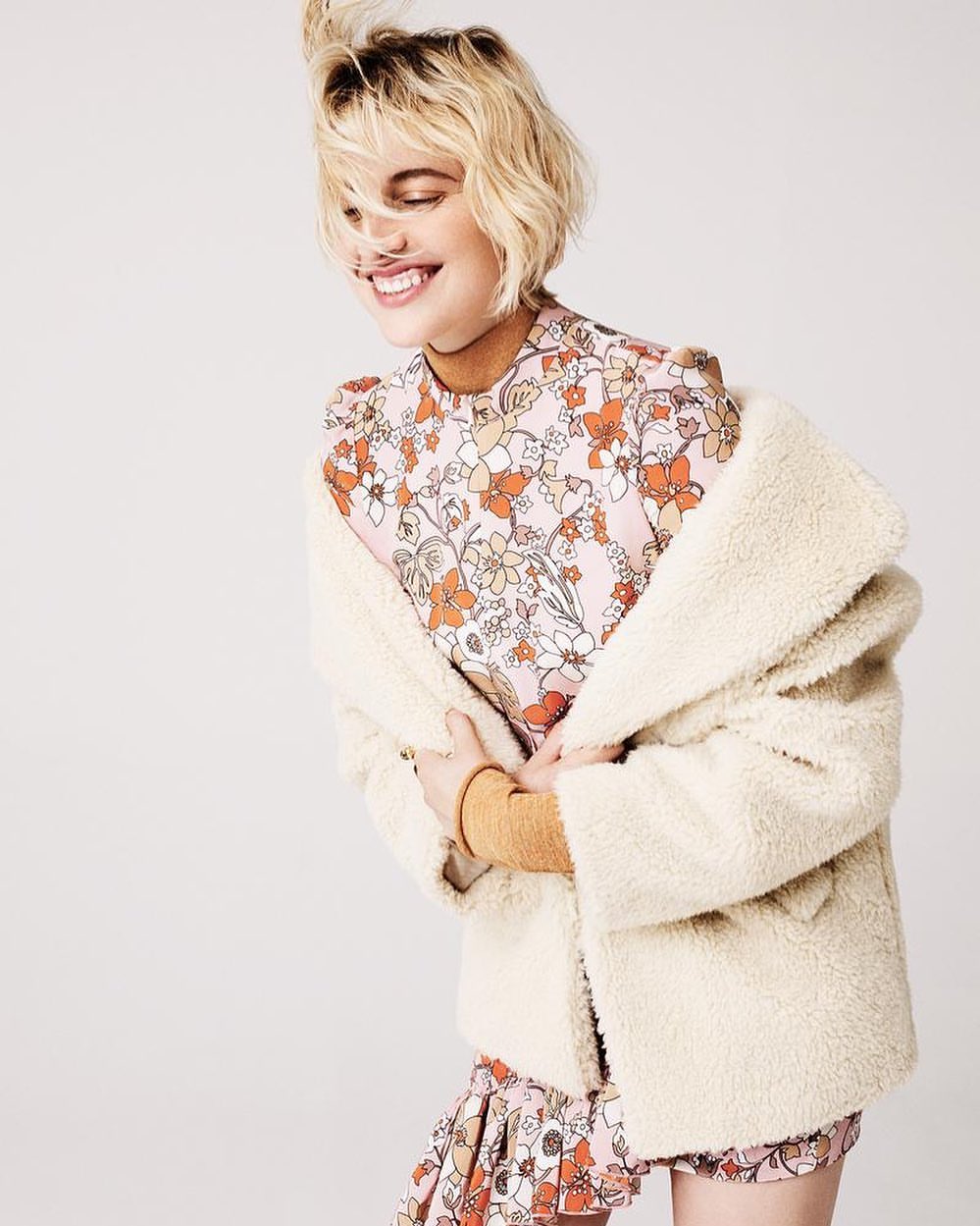 Smiling woman with short blonde hair in a floral print dress and a cream-colored sherpa jacket in a studio setting.