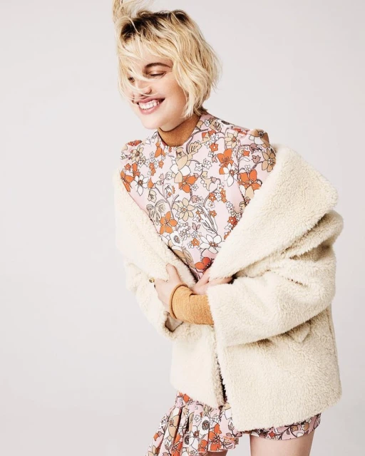 Smiling woman with short blonde hair in a floral print dress and a cream-colored sherpa jacket in a studio setting.