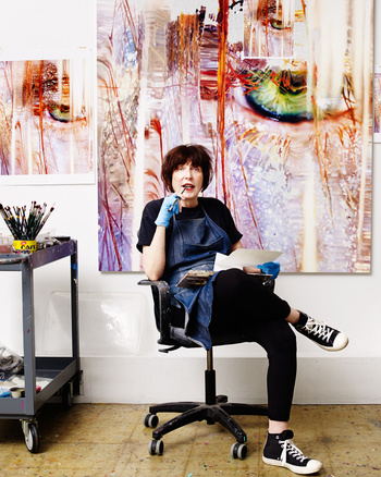 An artist sits in a rolling chair in her studio, with a large abstract painting behind her and painting supplies nearby.