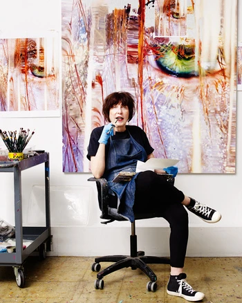 An artist sits in a rolling chair in her studio, with a large abstract painting behind her and painting supplies nearby.