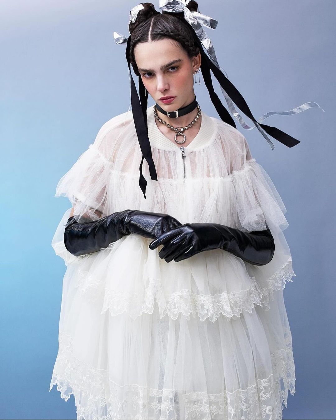 Young woman in white tulle dress and long black gloves poses against a blue background, accentuated by ribbons in her braided hair.