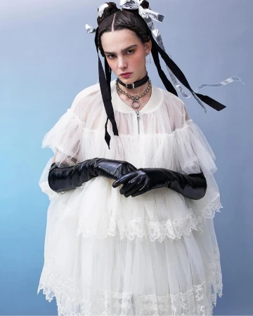 Young woman in white tulle dress and long black gloves poses against a blue background, accentuated by ribbons in her braided hair.