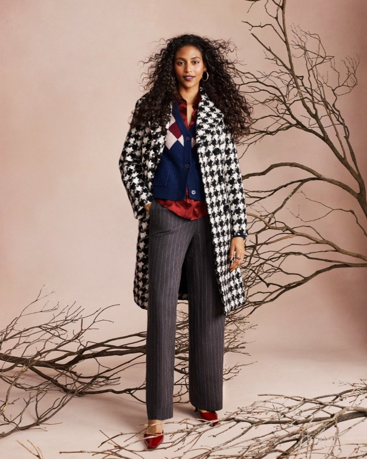 A woman in a houndstooth coat, red blouse, and striped pants stands in front of bare tree branches against a light pink backdrop.