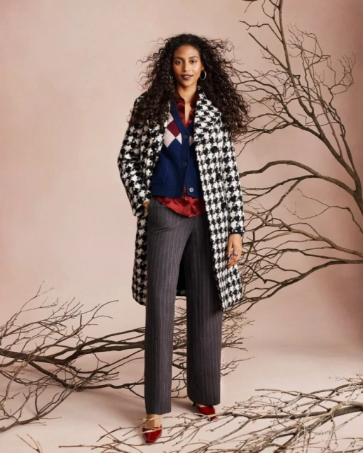 A woman in a houndstooth coat, red blouse, and striped pants stands in front of bare tree branches against a light pink backdrop.