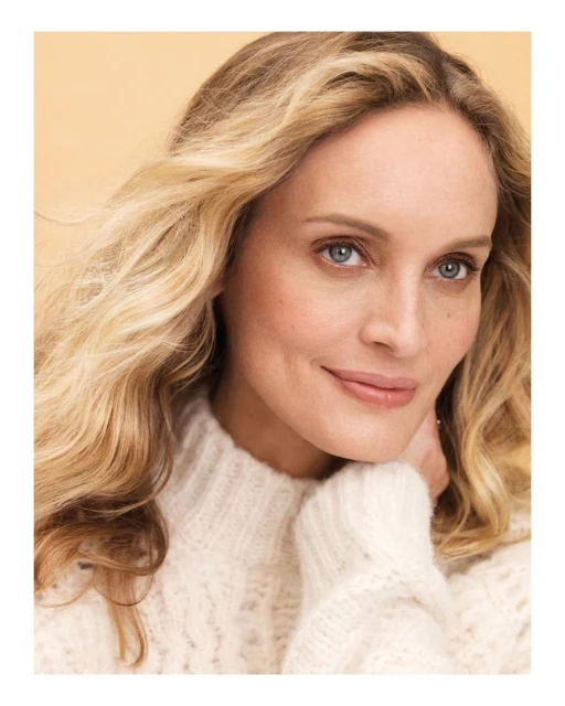 Blonde woman with wavy hair in a cream sweater poses for a portrait indoors with a peach background, smiling softly at the camera.