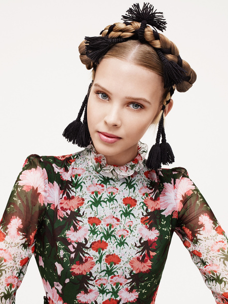Close-up portrait of a blonde adult female with braided hair, flower-patterned dress, and fair skin against a neutral background.