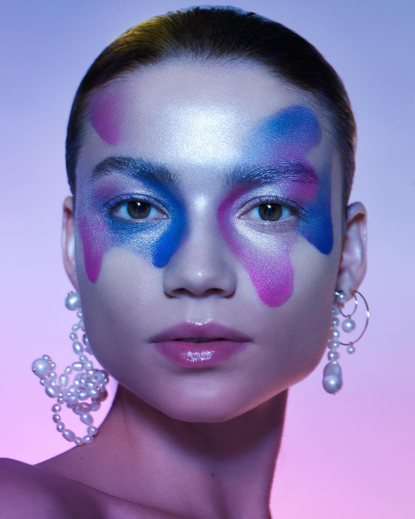 Close-up beauty shot of a woman with colorful face makeup and pearl earrings, set against a graduated purple and blue background.