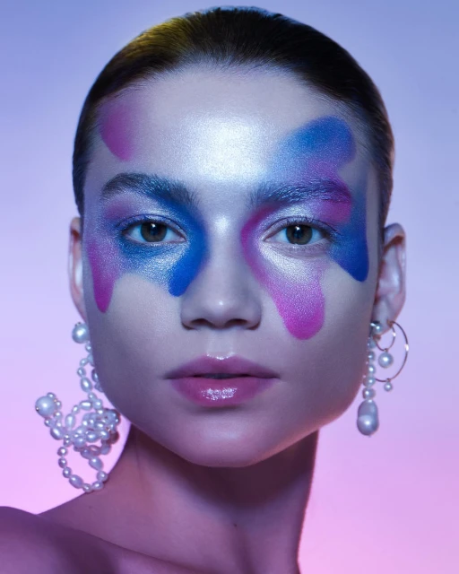 Close-up beauty shot of a woman with colorful face makeup and pearl earrings, set against a graduated purple and blue background.