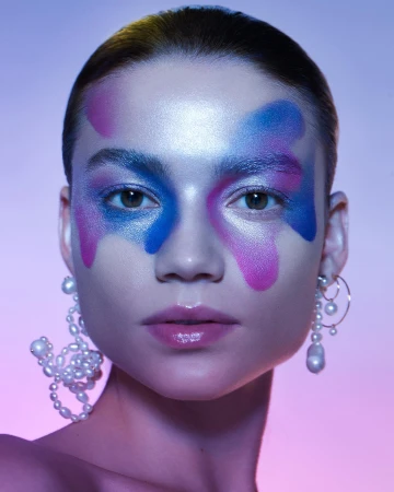 Close-up beauty shot of a woman with colorful face makeup and pearl earrings, set against a graduated purple and blue background.
